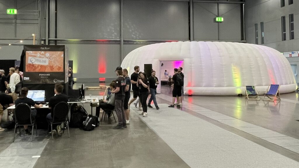 Große weiße, aufblasbare Kuppel in einer Messehalle, innen farbig ausgeleuchtet. Davor ein Spielstand mit Bildschirm und mehrere Besucher auf dem grauen Hallenboden.