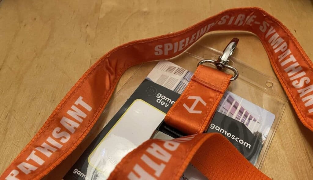 An orange trade fair lanyard printed with "Spieleindustrie-Sympathisant", lying flat on pale wood.